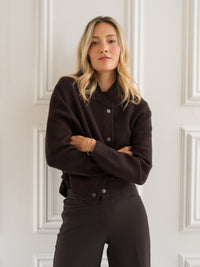 Brown bomber jacket in "Pure Choco" from Plein Publique, shown worn with arms crossed in front of a white paneled wall.
