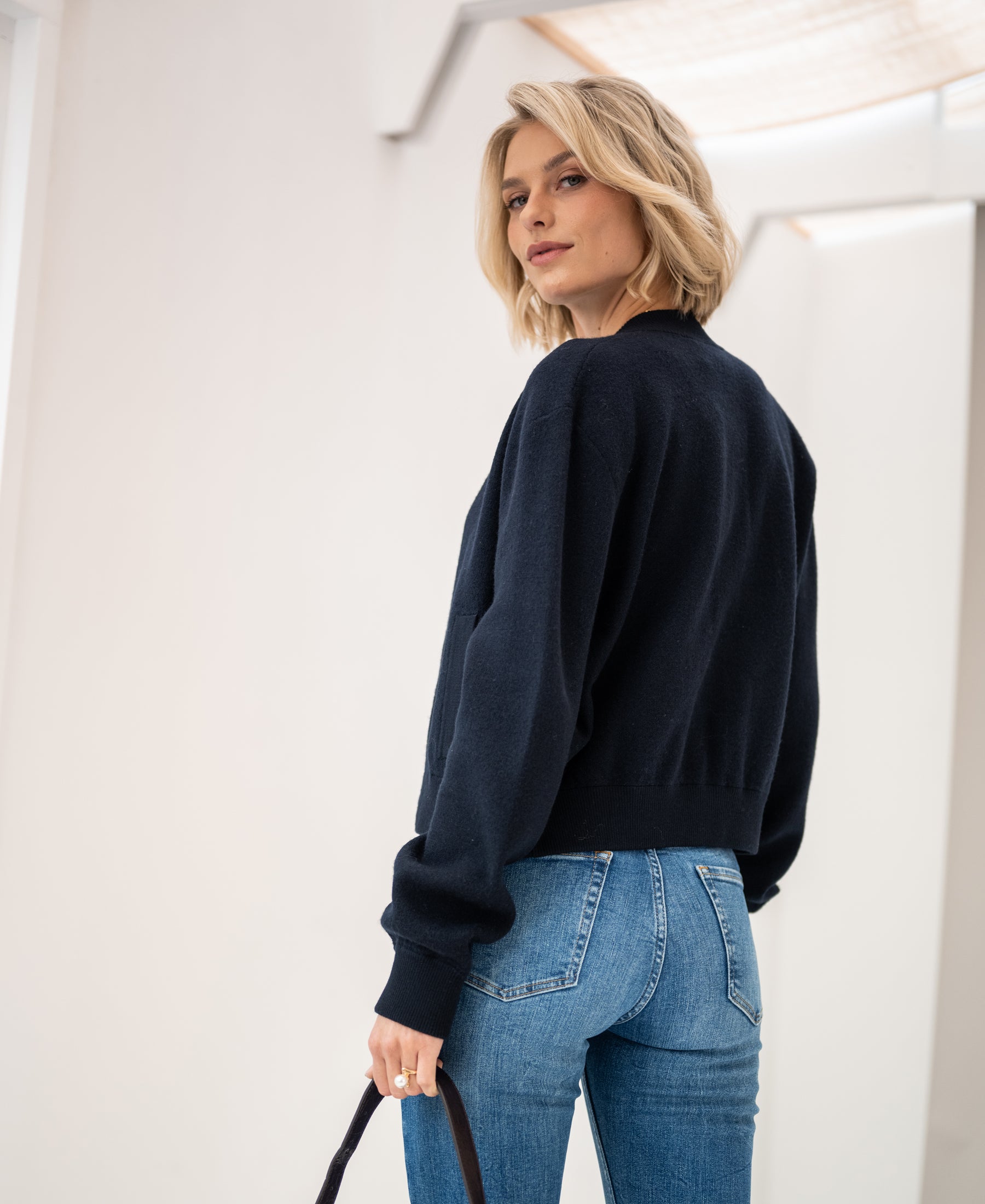 A dark blue bomber jacket, shown from the back as worn by a woman looking over her shoulder indoors.