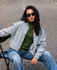 Woman wearing an olive LE FABIAN turtleneck sweater by PLEIN PUBLIQUE sits outdoors, looking at the camera.