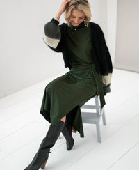Woman wearing an army green travel dress sits smiling on a gray chair.