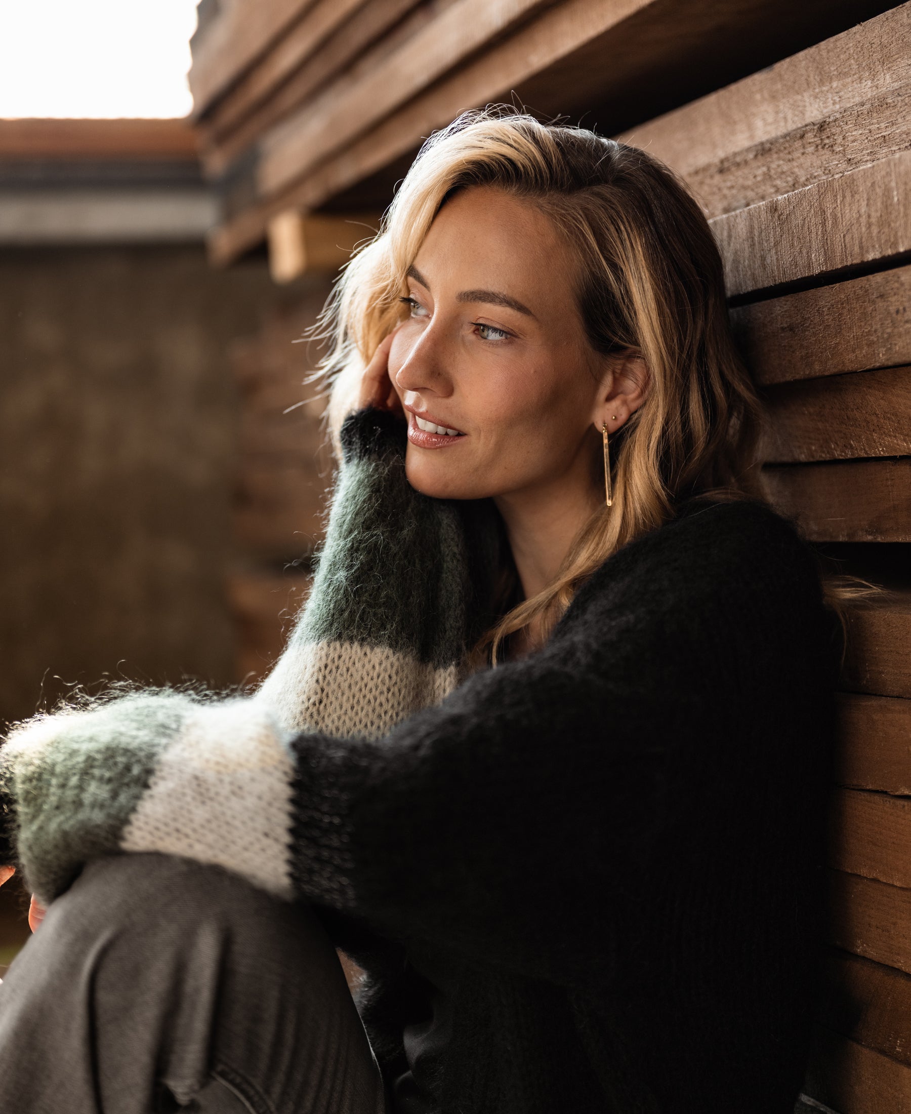 Woman wearing black and striped fluffy vest sits by a wooden wall.