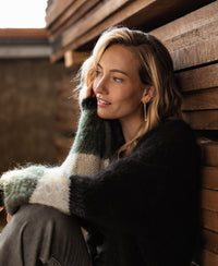 Woman wearing black and striped fluffy vest sits by a wooden wall.