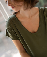Woman wearing an olive green ribbed T-shirt sits by a window, her face turned slightly down.