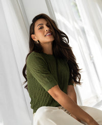 Woman wearing an olive green merino wool sweater stands by white curtains.