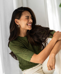 Woman wearing an olive green merino wool sweater, standing by a bright window.