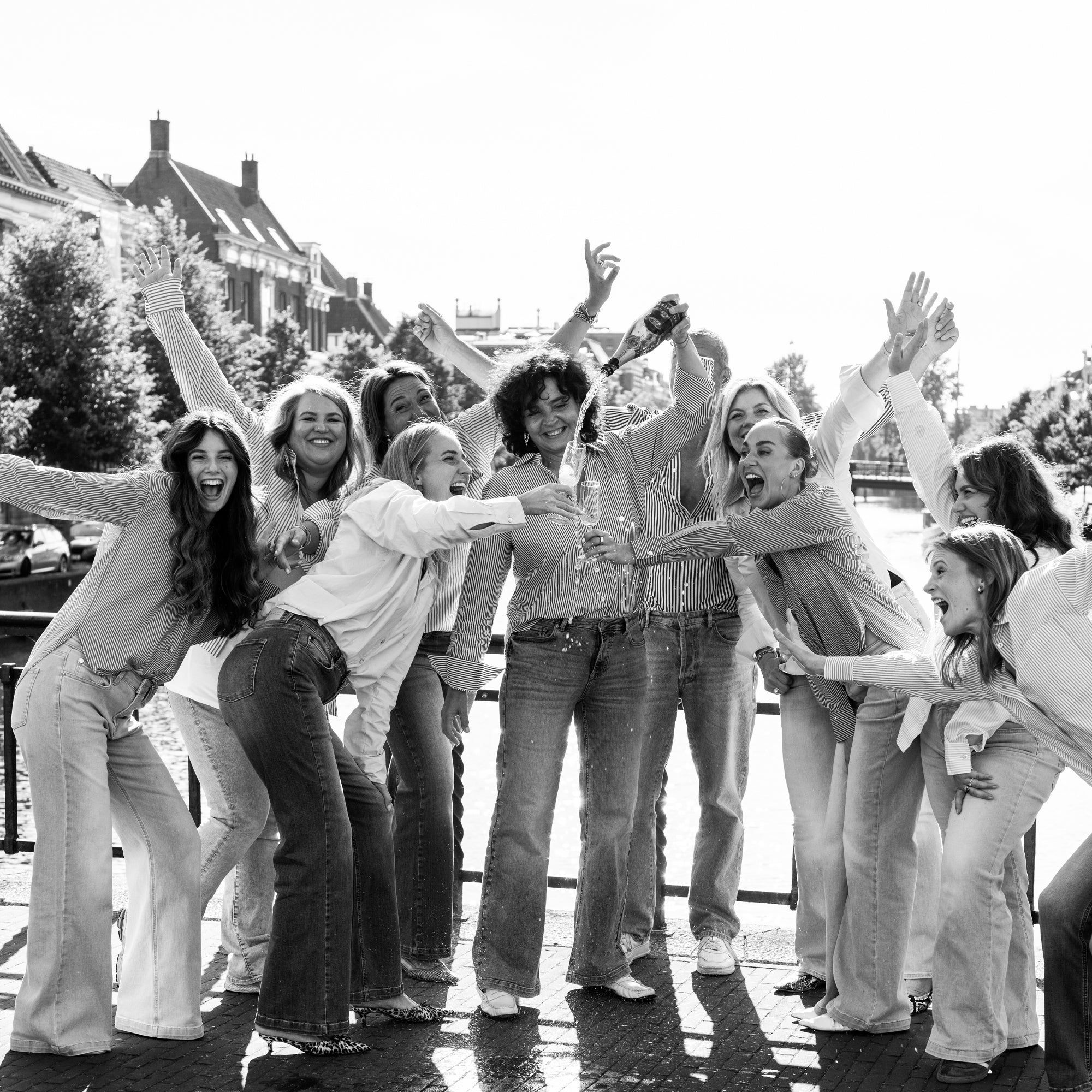 Women wearing flared jeans smile and laugh together outdoors by a canal on a sunny day.