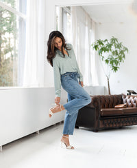 Light stone cropped jeans in denim, modeled indoors near a brown leather sofa and windows.