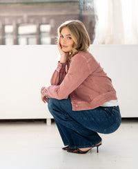 Woman wearing a dusty pink stretch denim jacket squats indoors, looking at the camera.