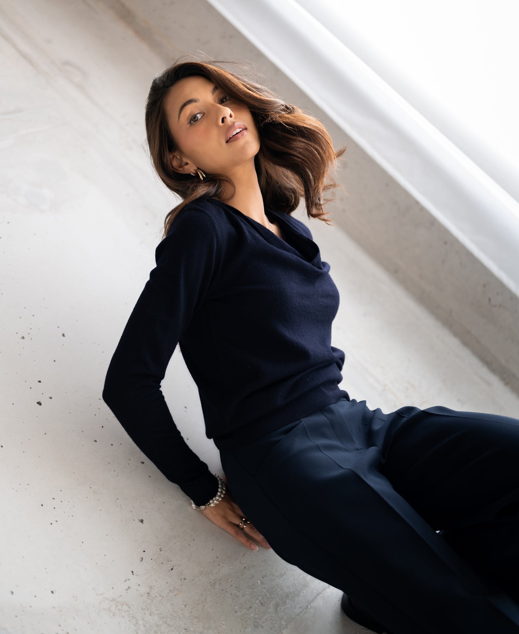Woman wearing PLEIN PUBLIQUE's LA CASCADE dark blue sweater, leaning back and looking confidently at the camera.