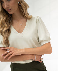 Stone-colored LA CELINE Traveltop by OVERIG with puff sleeves, worn by model, arms crossed, blurred background.
