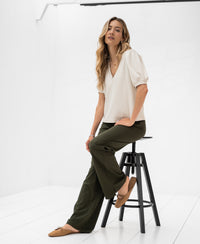 White women’s travel top in stone color shown on a model seated in a bright, minimalist room.