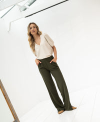 Woman wearing a gray OVERIG travel top, standing in a minimal, bright room with hands in pockets.