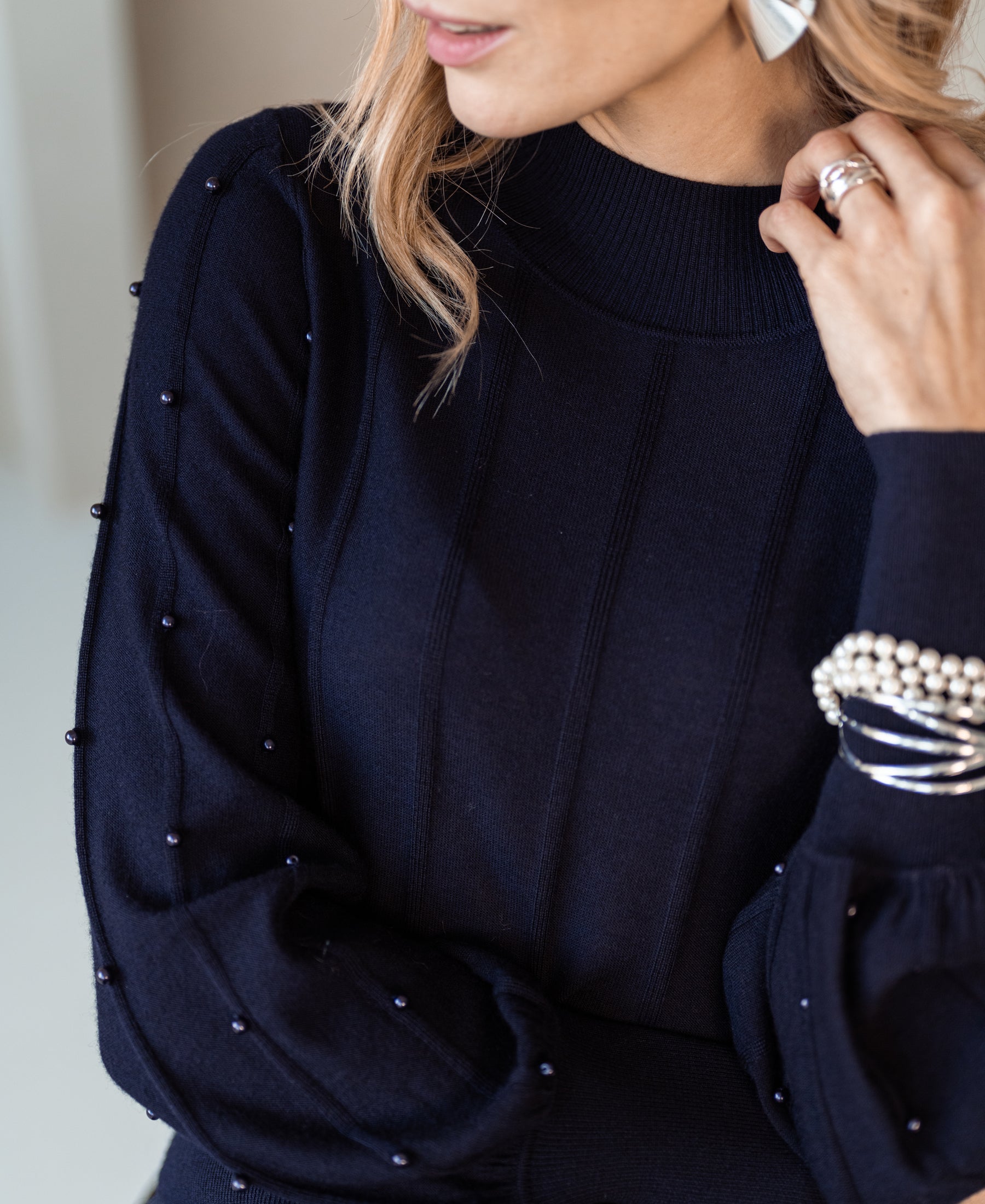 Woman wearing PLEIN PUBLIQUE LA CENTAINE dark blue turtleneck sweater with beads, touching her face.