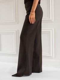 Wide-leg chocolate brown pants from Plein Publique shown against a white paneled wall.