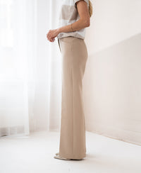 Wide-leg light sand trousers, side view, indoors, white curtains in background.
