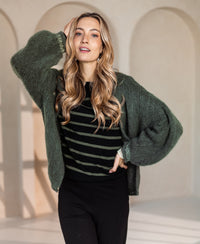 Woman poses indoors in a black and army green PLEIN PUBLIQUE L'ELISA dress, one hand on hip, the other by her head.