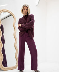 Woman wearing fluffy Bordeaux vest, standing by a wavy mirror in a minimalist room.