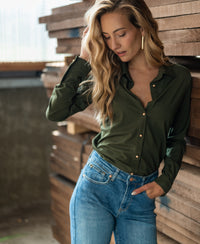 Woman wearing the PLEIN PUBLIQUE LA CHERISE Army green travel blouse stands by stacked wood, touching her wavy hair.