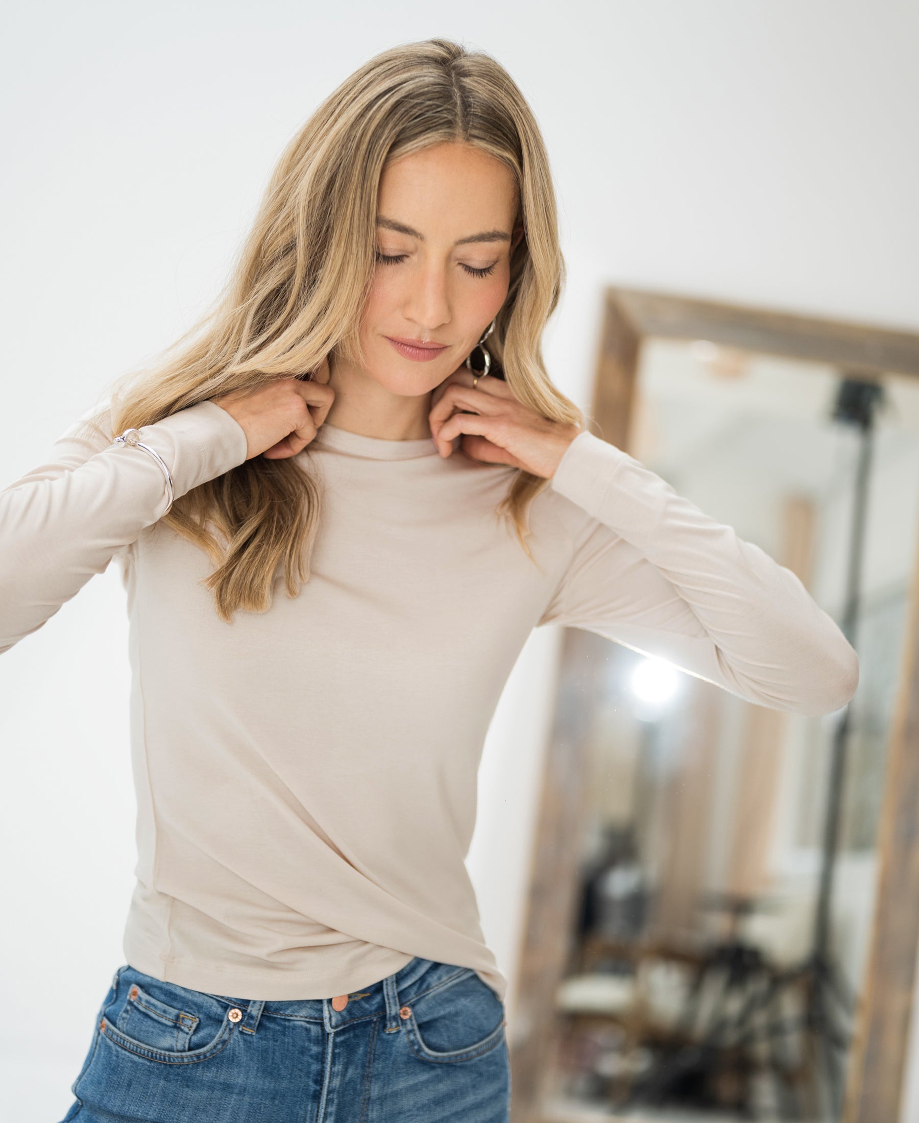 A woman adjusts her Luxe undershirt LE CHOLET light sand by PLEIN PUBLIQUE in front of a mirror in a bright room.