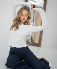 Woman wears the PLEIN PUBLIQUE Luxe undershirt LE CHOLET White, seated on the floor with arms raised in a bright room.