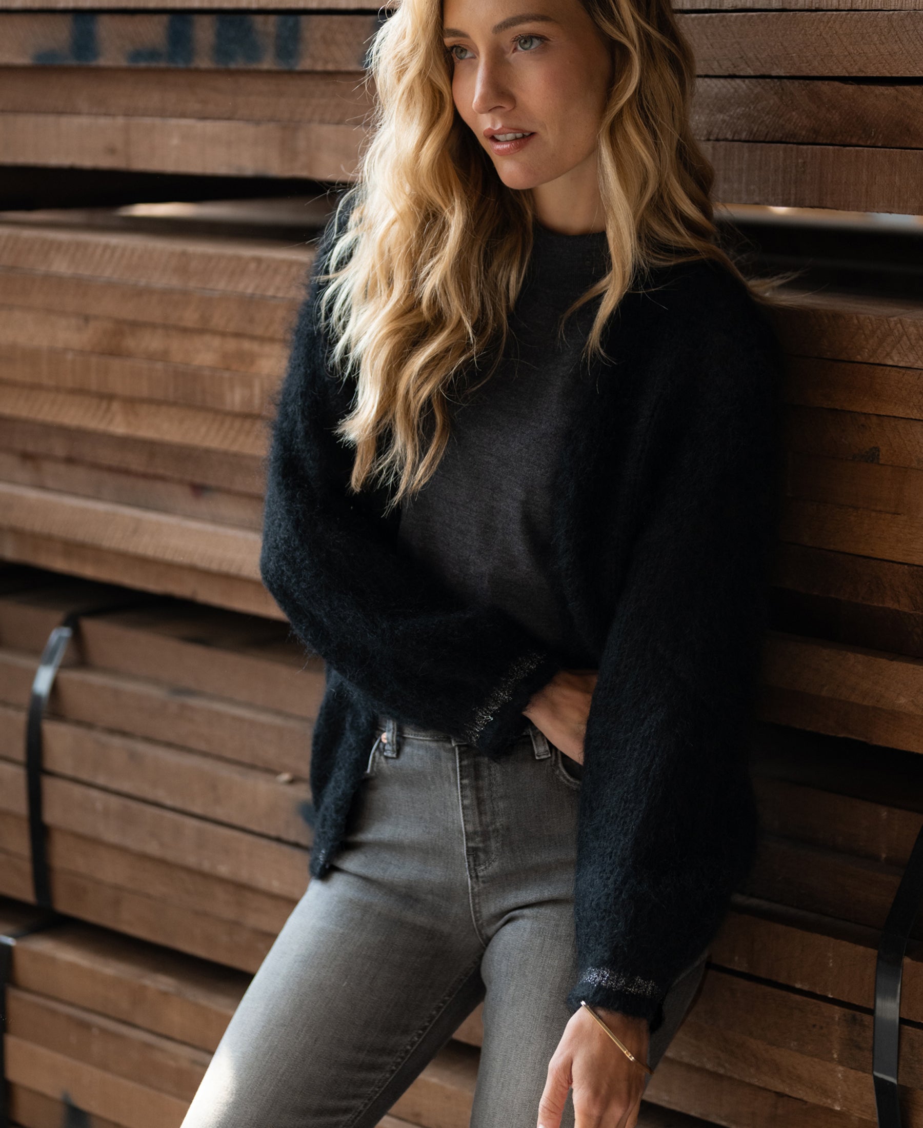 Woman wearing the PLEIN PUBLIQUE Fluffy black vest leans on stacked wooden planks.