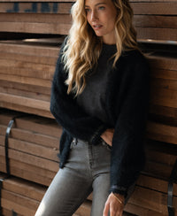 Woman wearing the PLEIN PUBLIQUE Fluffy black vest leans on stacked wooden planks.