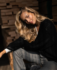 Woman in sunlight wears the black PLEIN PUBLIQUE LA CHERIE fluffy vest by stacked wood.