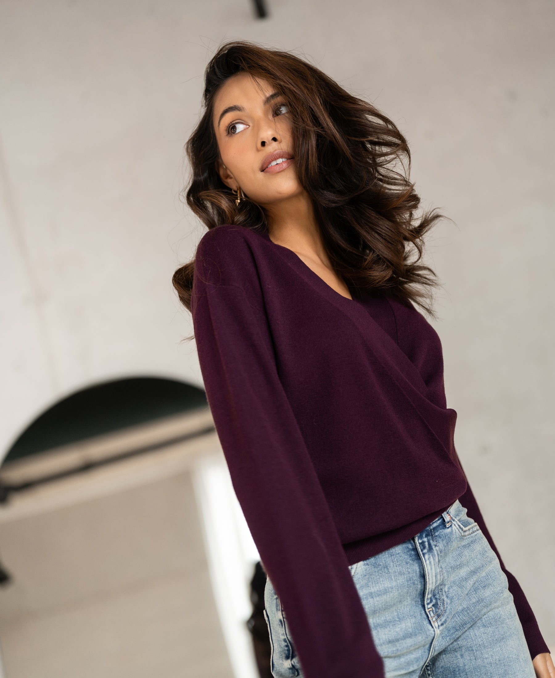 Woman wearing a PLEIN PUBLIQUE wrap sweater in aubergine stands indoors, shown from the front.