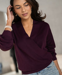 Woman wearing the LES CINQ eggplant wrap sweater by PLEIN PUBLIQUE, touching her hair and smiling.