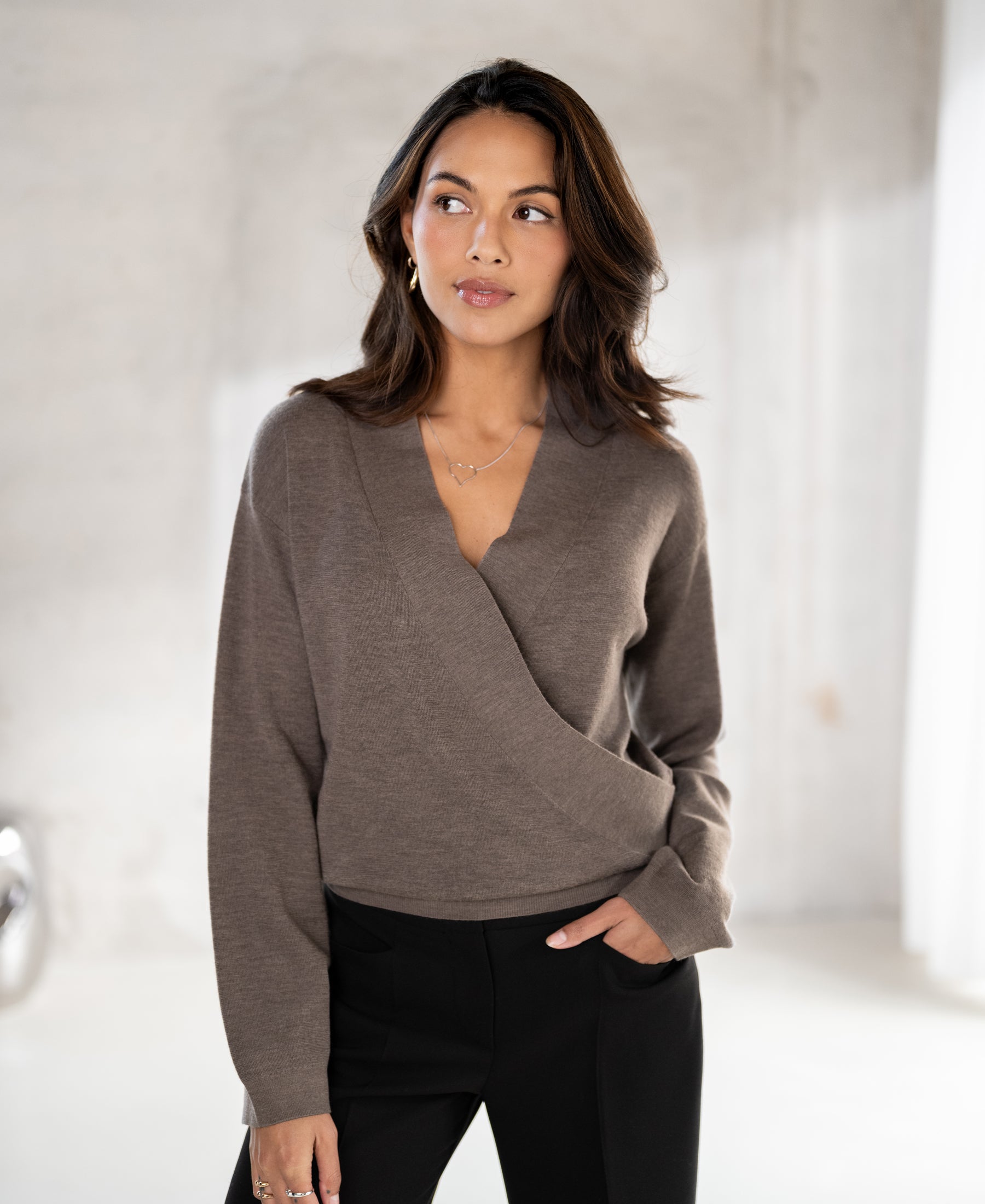 A woman with wavy dark hair wears the PLEIN PUBLIQUE wrap sweater in taupe melange indoors.