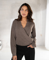 A woman with wavy dark hair wears the PLEIN PUBLIQUE wrap sweater in taupe melange indoors.