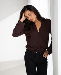 Woman wearing the LES CINQ Cacao wrap sweater by PLEIN PUBLIQUE stands by a curtain, eyes closed, hand resting on her head.