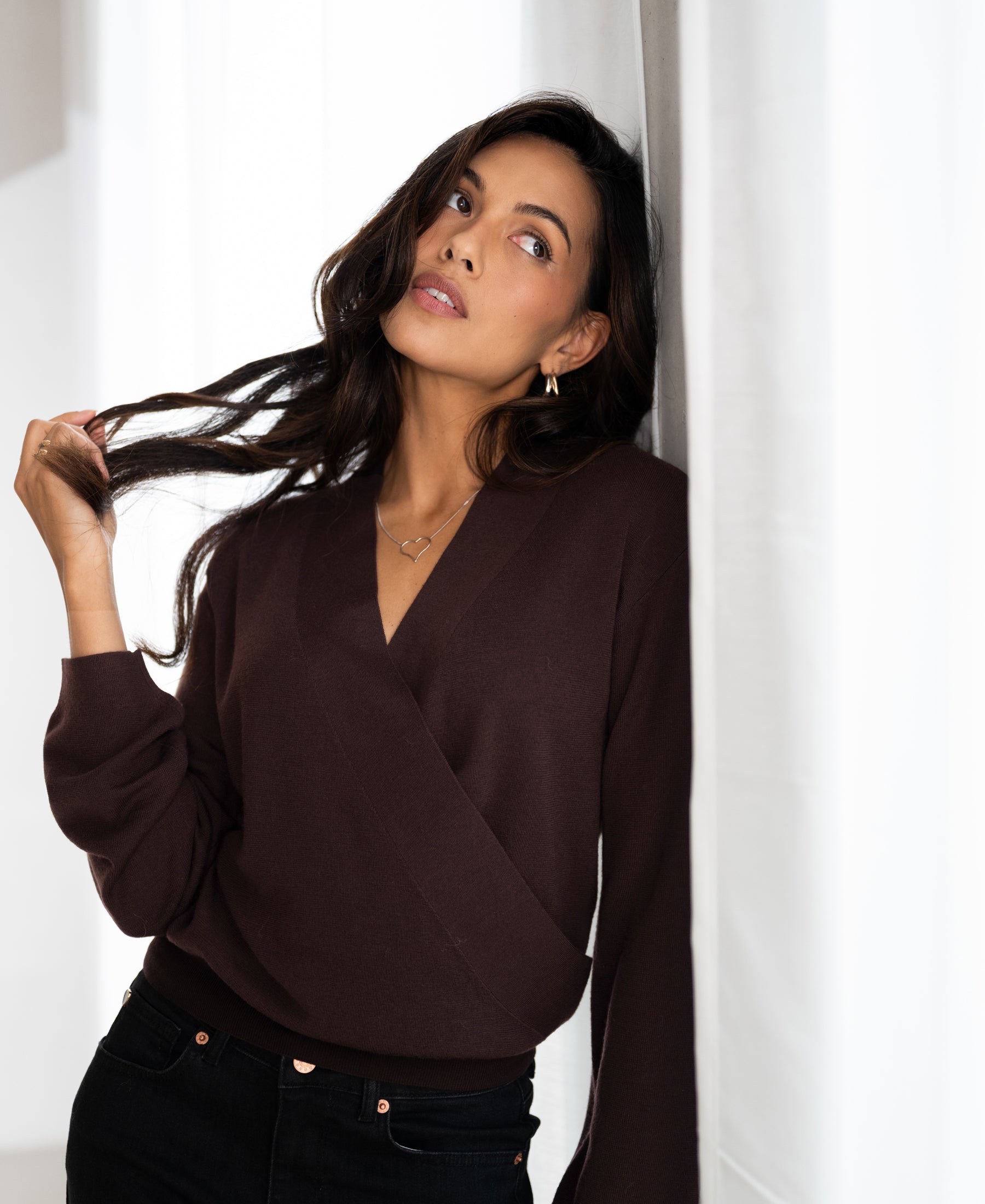 Woman wearing the LES CINQ Cacao wrap sweater by PLEIN PUBLIQUE leans on a white wall, looking up and touching her hair.