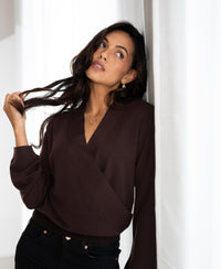 Woman wearing the LES CINQ Cacao wrap sweater by PLEIN PUBLIQUE leans on a white wall, looking up and touching her hair.