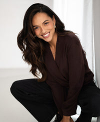 A woman wears a finely knitted wrap sweater in cacao brown by PLEIN PUBLIQUE indoors near white curtains.