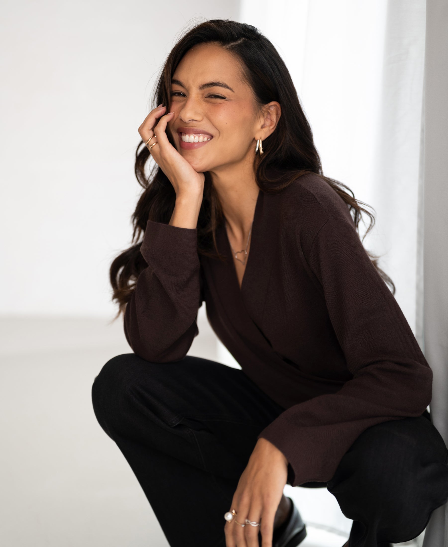 Woman wearing the PLEIN PUBLIQUE wrap sweater LES CINQ in cacao, hand on cheek, crouching.