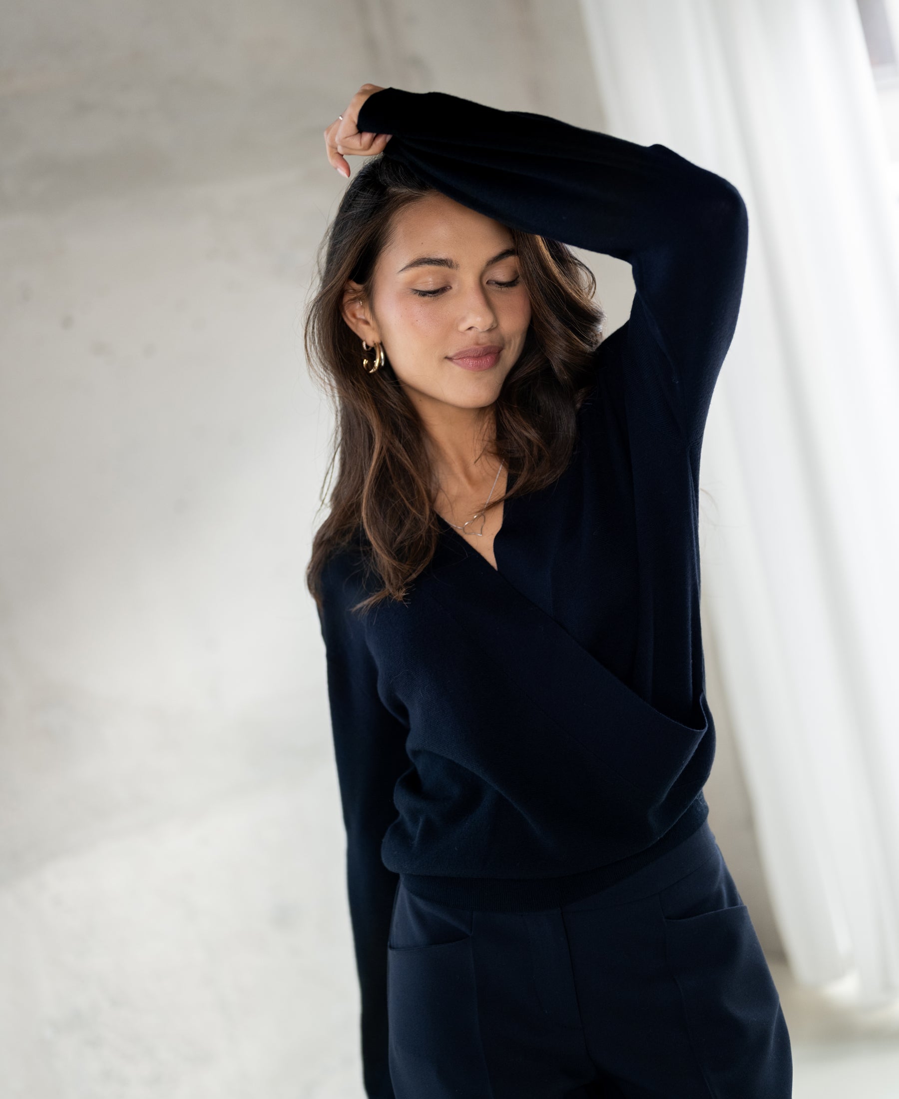 Donkerblauwe LES CINQ wrap sweater van PLEIN PUBLIQUE getoond bij zacht licht voor witte gordijnen.