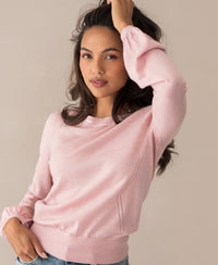 A woman with long, wavy brown hair wears the Merinotrui LA COEUR Rose and light blue jeans, posing confidently with one hand in her hair against a neutral beige background.