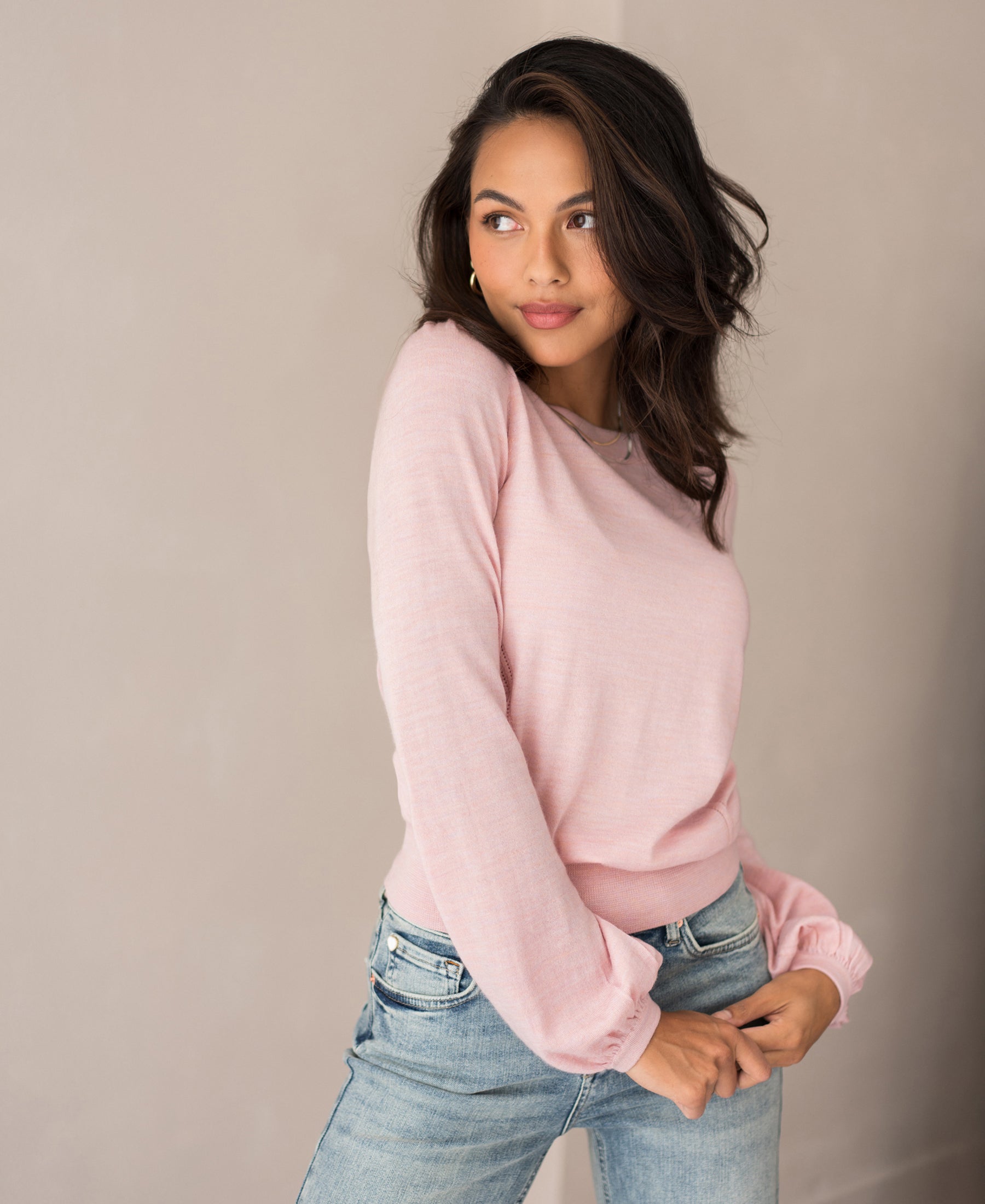 A woman with long dark hair wears the Merinotrui LA COEUR Rose, a light pink merino wool sweater with ajour details, paired with blue jeans. She stands against a plain light background, gazing gently to her left.