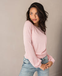 A woman with long dark hair wears the Merinotrui LA COEUR Rose, a light pink merino wool sweater with ajour details, paired with blue jeans. She stands against a plain light background, gazing gently to her left.