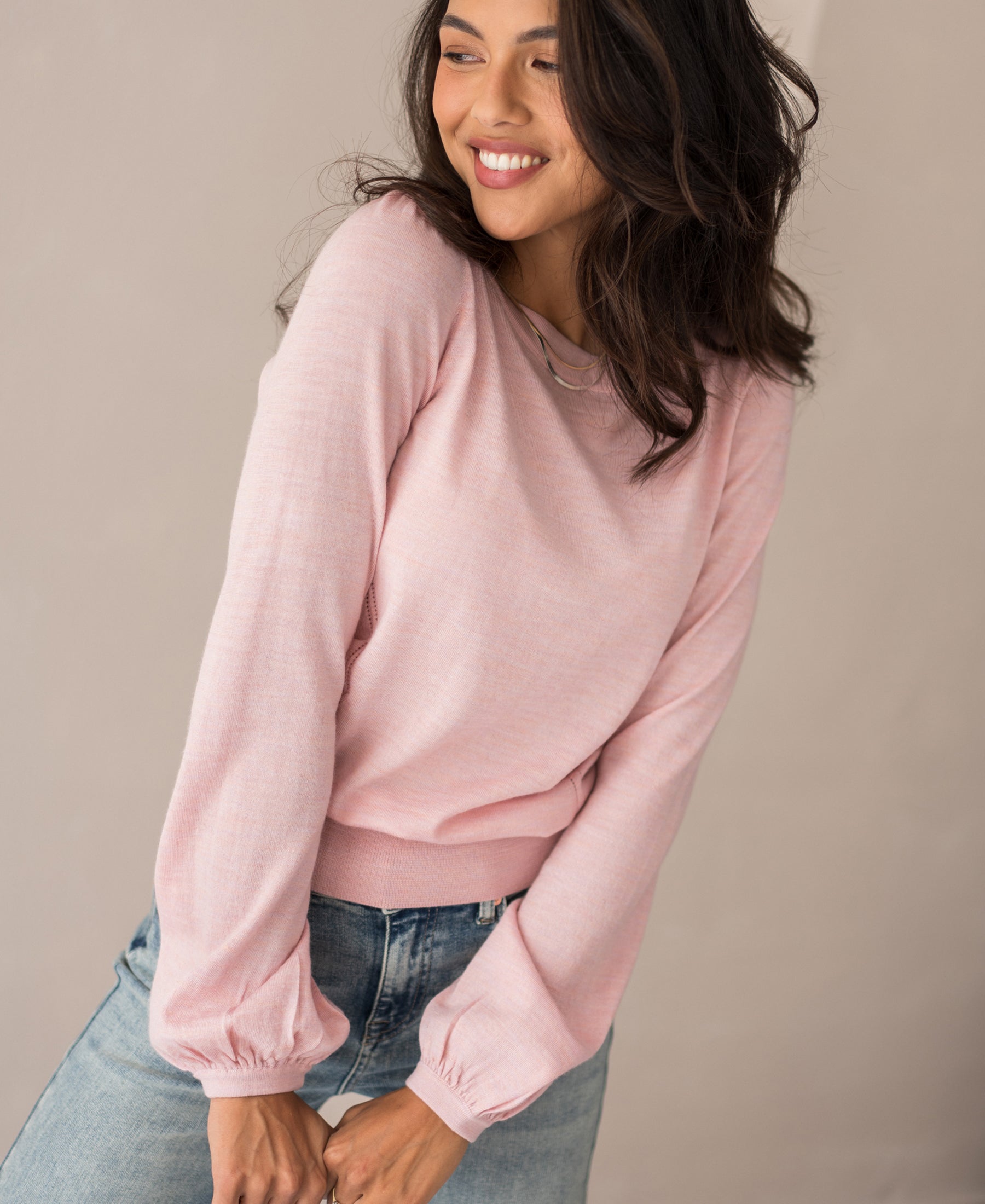 A woman with wavy dark hair smiles while looking to the side, wearing the Merinotrui LA COEUR Rose and blue jeans, standing against a plain, neutral background.
