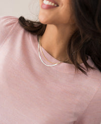 A woman with wavy dark hair smiles, wearing the Merinotrui LA COEUR Rose—a light pink top with balloon sleeves—and two delicate layered gold necklaces.