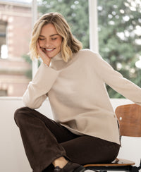 Woman wearing a beige LA COZY turtleneck sweater sits indoors, resting her chin on her hand.