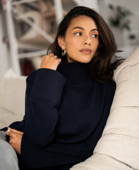 Woman wearing the LA COZY dark blue turtleneck sweater by PLEIN PUBLIQUE, seated on a light sofa, looking to the side.