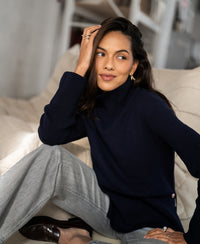 Woman wearing a PLEIN PUBLIQUE LA COZY dark blue turtleneck sweater sits on a cream couch, smiling.
