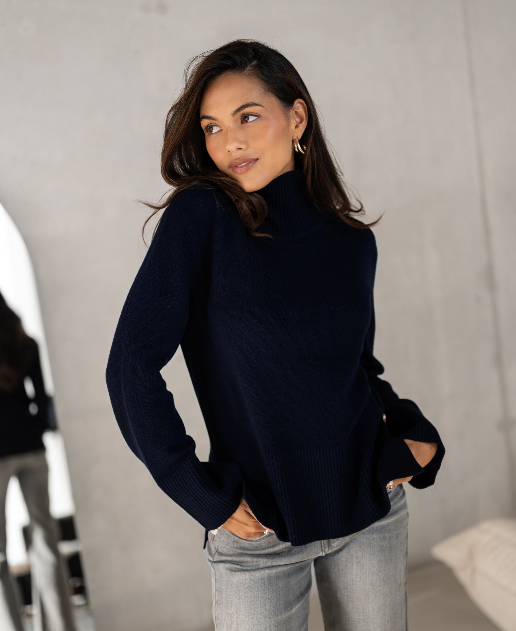 Woman wearing dark blue PLEIN PUBLIQUE LA COZY turtleneck sweater, standing indoors, looking to the side.