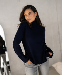 Woman wearing dark blue PLEIN PUBLIQUE LA COZY turtleneck sweater, standing indoors, looking to the side.