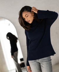 Woman wearing a PLEIN PUBLIQUE LA COZY dark blue turtleneck sweater stands indoors by a mirror.