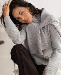 Woman wears the PLEIN PUBLIQUE LA COZY grey turtleneck sweater, sitting and looking at camera, touching her hair.