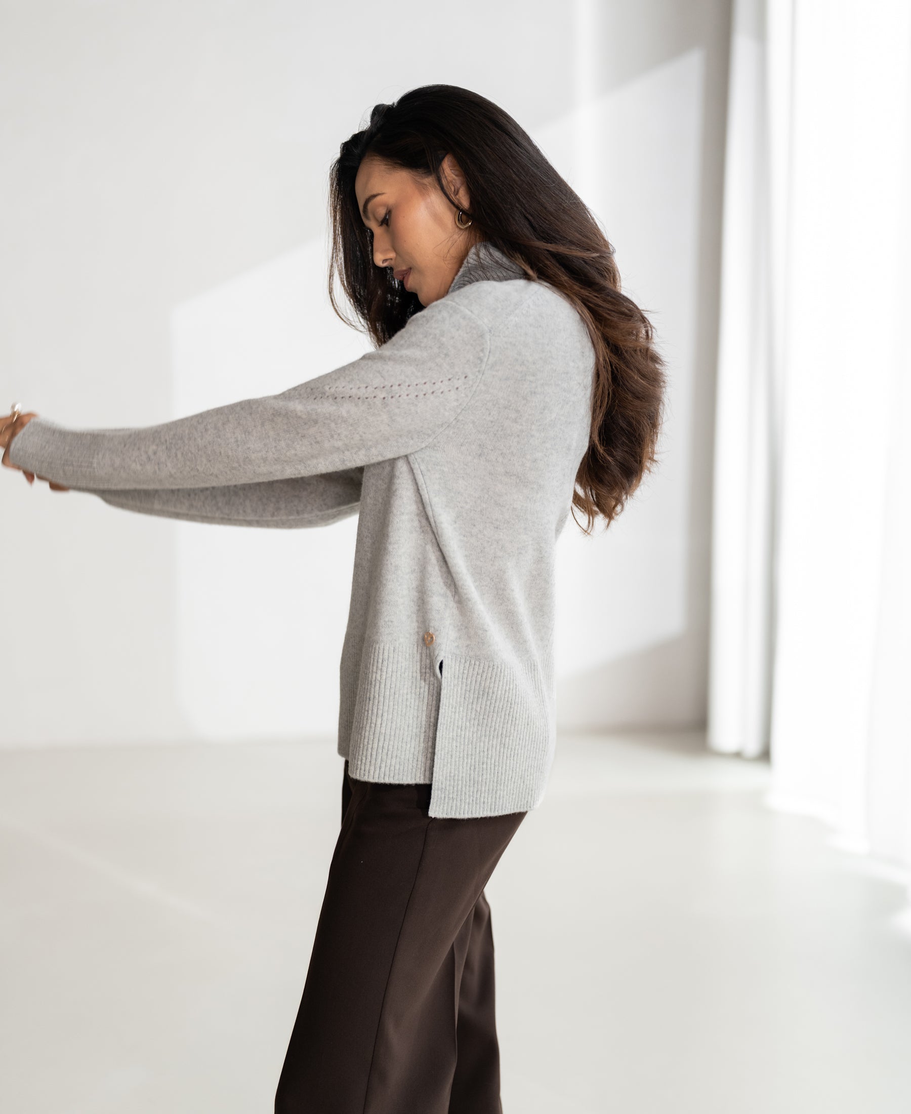 Woman wearing a grey LA COZY turtleneck sweater stands in a bright room with white curtains.